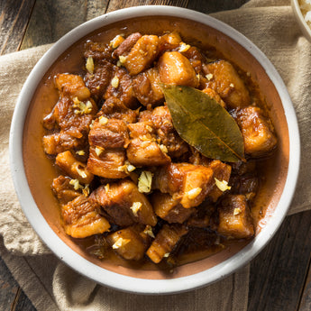 A bowl of pork adobo (a Filipino dish) with vegetables, served on a wooden table with a white napkin beside it.