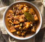 A bowl of pork adobo (a Filipino dish) with vegetables, served on a wooden table with a white napkin beside it.